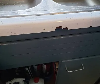 Damaged white sink edge above dark gray cabinet with cleaning supplies visible.