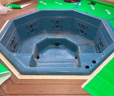 Blue hot tub with built-in seats and jets, surrounded by wooden decking and green protective covers.