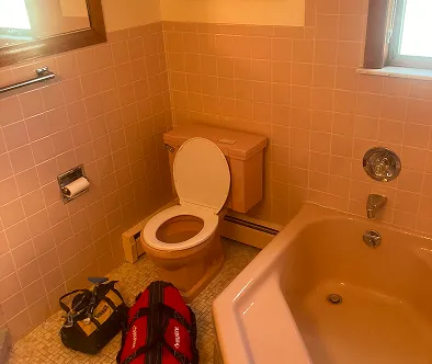 Bathroom with pink tiled walls, toilet, bathtub, and bags on the floor.