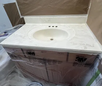 White bathroom vanity with a sink and faucet, wrapped in plastic and cardboard, with a brown wall behind it.